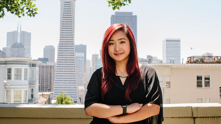 Student leans against a railing with the city skyline behind.