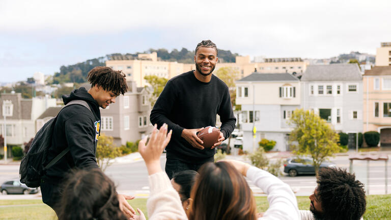 student holds football while working towards a group of other students sitting on the lawn
